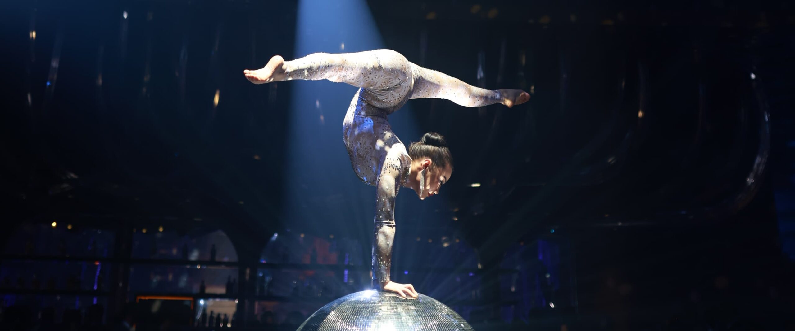 Aerial acrobat performing a one-hand balance pose on a mirrored sphere under a spotlight during a live stage performance.