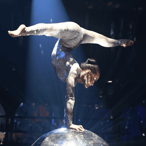 Aerial acrobat performing a one-hand balance pose on a mirrored sphere under a spotlight during a live stage performance.